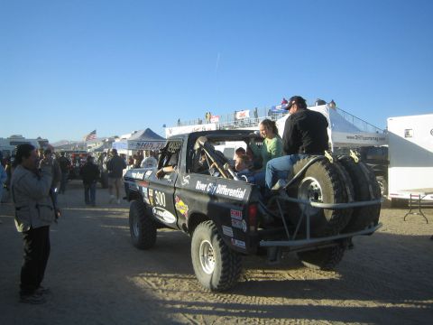 1990 Ford Bronco - Rear of black race truck number 300 with a mounted spare tire and people aboard in a busy desert pit area.