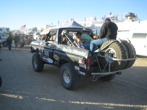1990 Ford Bronco - black off-road race truck marked 300 and "River City Differential" sits in a dusty event pit with crew and tents in the background.