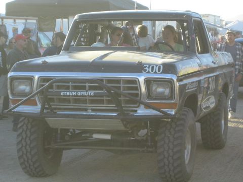 1990 Ford Bronco - front end of a black off-road race truck numbered 300 shows a tubular brush guard, big knobby tires and a lifted suspension among onlookers.
