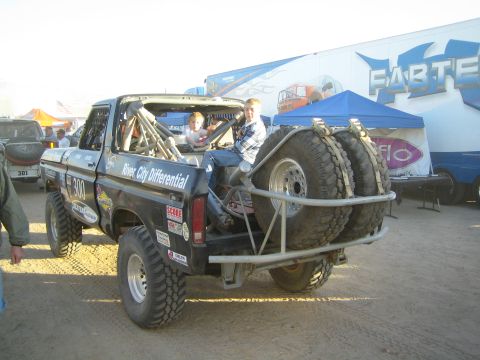 1990 Ford Bronco - Two boys stand in the bed of a black off-road race truck carrying twin rear spare tires in a busy event pit with team trailers behind.