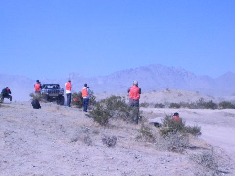 1990 Ford Bronco - Spectators in orange safety vests stand on a desert ridge photographing a dark truck on the course with rugged mountains in the distance.