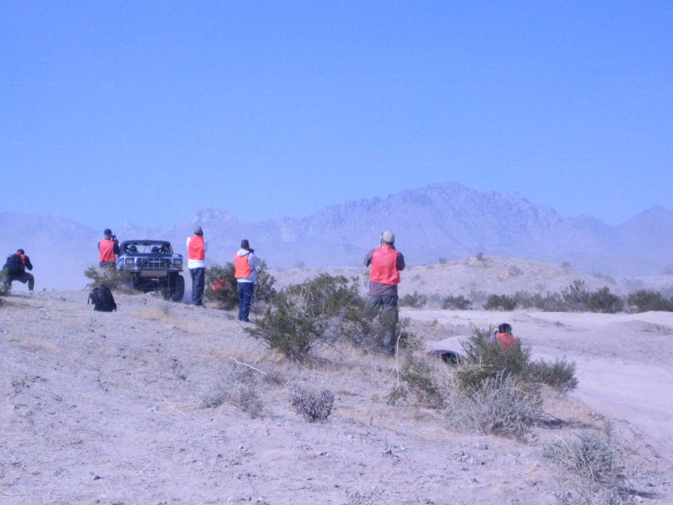 People in orange vests watch from a desert hill as a truck waits below