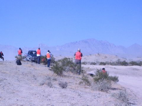 1990 Ford Bronco - group of people in orange vests watch and photograph a dark race truck on a dusty desert course backed by distant mountains.