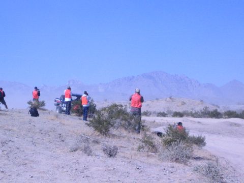 1990 Ford Bronco - People in orange safety vests gather on a brushy desert hill beside a dark vehicle, watching the course under a clear blue sky.