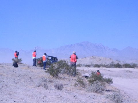 1990 Ford Bronco - Onlookers in orange vests stand on a sandy desert rise watching a dark vehicle, with a hazy mountain ridge along the horizon.