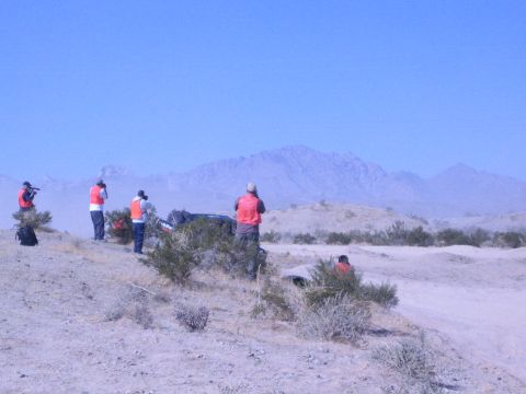 1990 Ford Bronco - cluster of people in orange vests photograph a dark vehicle across a dusty desert wash framed by distant mountains.