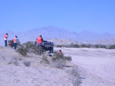 1990 Ford Bronco - dark lifted truck sits on a sandy desert course as orange-vested spectators look on beneath rugged blue mountains.