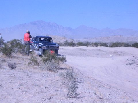 1990 Ford Bronco - black lifted race truck waits on a sandy desert trail while a lone spectator in an orange vest stands nearby below distant peaks.