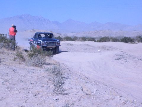 1990 Ford Bronco - black lifted race truck rolls along a dusty desert course as an orange-vested spectator photographs it with mountains beyond.