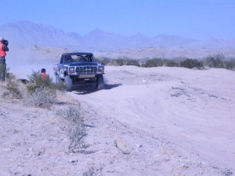 1990 Ford Bronco - black lifted off-road race truck climbs a sandy berm on a dusty desert course with spectators at the trailside.