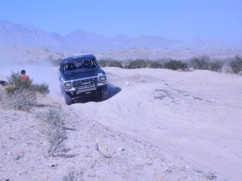 1990 Ford Bronco - black lifted race truck numbered 300 powers through deep sand on a desert course, throwing up a trail of dust.