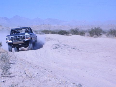1990 Ford Bronco - black off-road race truck marked 300 charges across a sandy desert wash, kicking up a plume of dust under a clear sky.