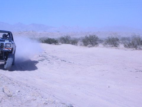 1990 Ford Bronco - black lifted race truck numbered 300 speeds along a sandy desert track, trailing dust beside distant mountains and power lines.