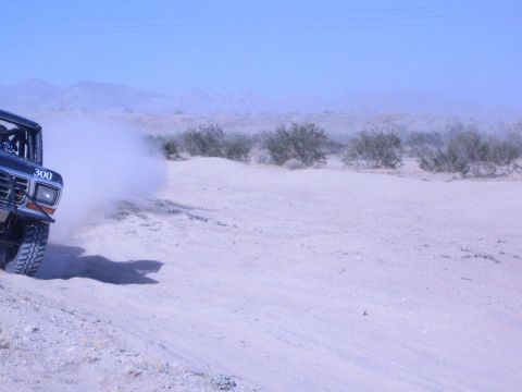 1990 Ford Bronco - black off-road race truck marked 300 sprays a wall of sand as it powers through a dusty desert course.