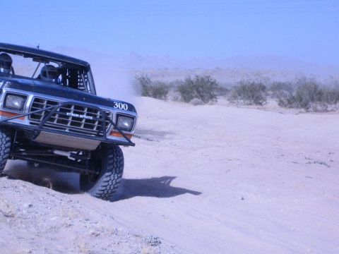 1990 Ford Bronco - close low-angle view of a black race truck numbered 300 shows its brush guard, big tires and lifted front end on a sandy course.