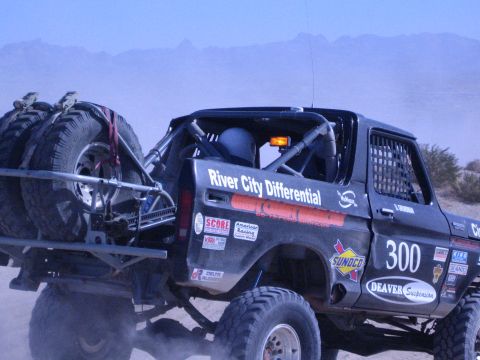 1990 Ford Bronco - rear of a black race truck numbered 300 with twin lashed spare tires kicks up dust as it races across the desert.