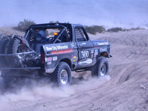1990 Ford Bronco - rear three-quarter view of black race truck 300 shows it powering through sand, trailing dust under hazy desert hills.