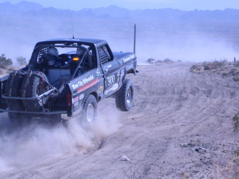 1990 Ford Bronco - Black off-road race truck 300 speeds along a dusty desert course as another vehicle follows in the distance.
