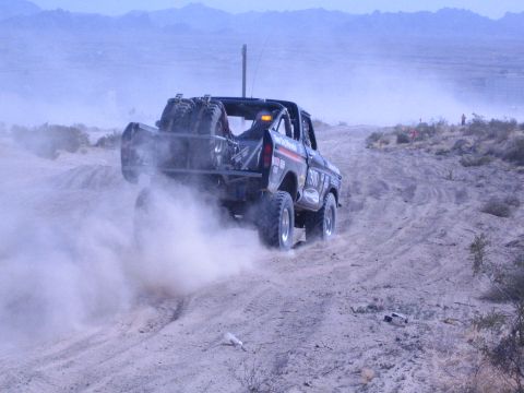 1990 Ford Bronco - black off-road race truck with spare tires mounted on its bed throws up a cloud of dust on a desert course.