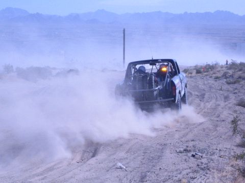 1990 Ford Bronco - dark off-road race truck races away through thick dust on a sandy desert trail with mountains in the distance.