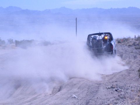 1990 Ford Bronco - off-road race truck recedes into a cloud of dust along a desert course under a hazy sky.