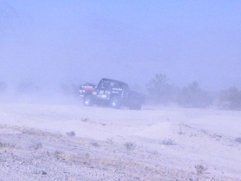 1990 Ford Bronco - black off-road race truck marked 300 with sponsor decals drives across open dusty desert.