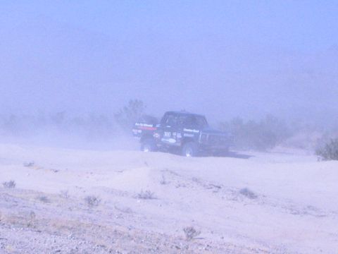 1990 Ford Bronco - off-road race truck numbered 300 crosses sandy desert terrain partly veiled in blowing dust.