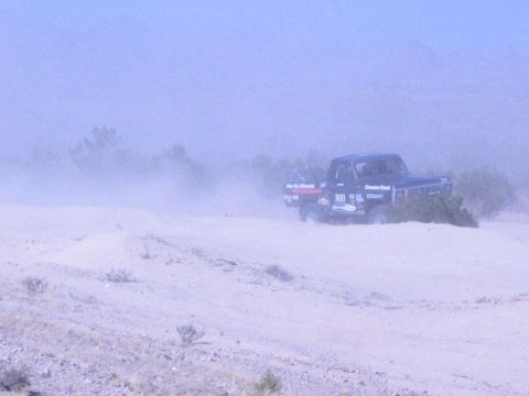 1990 Ford Bronco - black off-road race truck numbered 300 with sponsor decals drives past low desert brush in a dusty haze.
