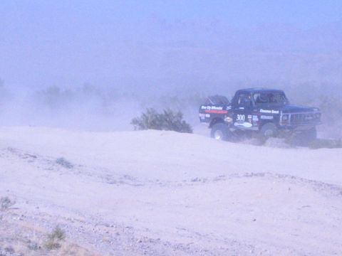 1990 Ford Bronco - off-road race truck numbered 300 climbs a sandy desert berm, trailing dust under a pale sky.