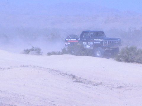 1990 Ford Bronco - black off-road race truck numbered 300 traverses a sandy desert ridge dotted with brush.