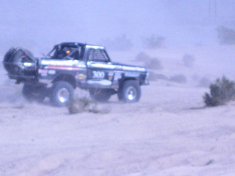 1990 Ford Bronco - black off-road race truck numbered 300 catches air over a desert rise, kicking up dust.