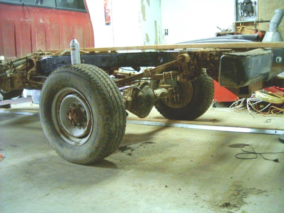 Stripped pickup frame with bed removed showing rusty rear axle, leaf springs and steel wheels