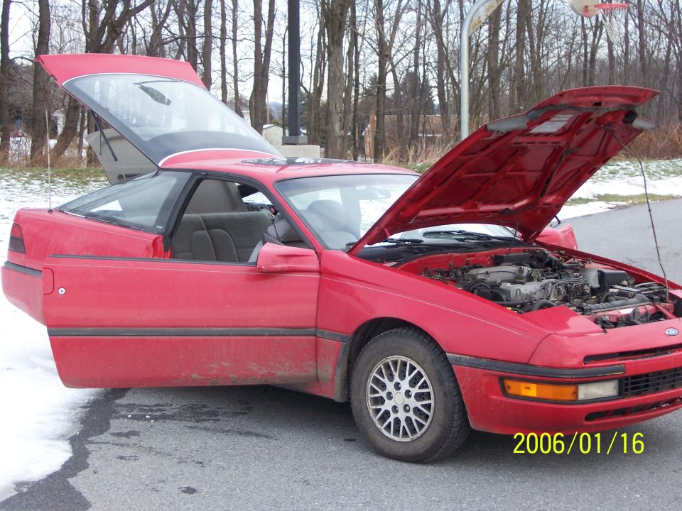 Red Ford Probe coupe with hood and hatch open in a parking lot, bare trees behind