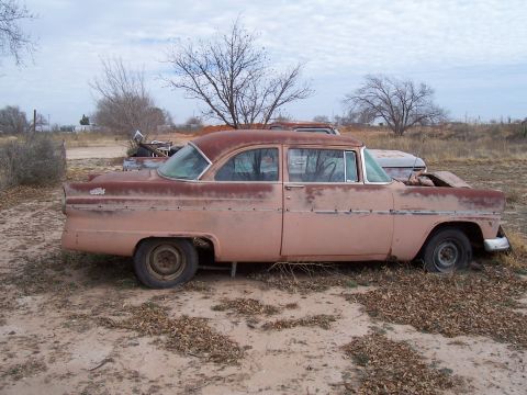 1955 Ford Customline - A weathered 1955 Ford Customline sedan sits in a dry, outdoor lot, showing its age with rust and faded paint.