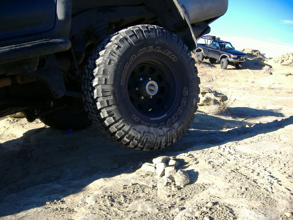 Close-up of a large off-road tire on a rocky trail, with another truck in the background