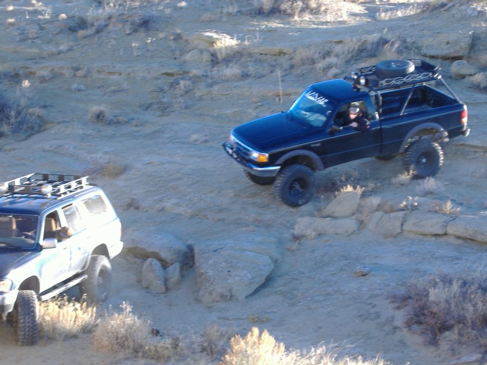 Black Ford Ranger pickup truck with a roof rack and spare tire, driving on a rocky incline