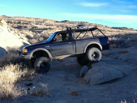 1978 Ford Bronco - Modified black Ford Ranger pickup truck driving over rocks in a dry, hilly landscape.