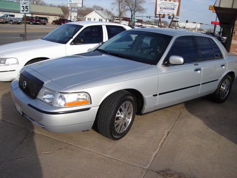 2000 Ford Crown Victoria - Two sedans, a silver and a white one, parked on a paved lot.
