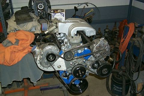 1978 Ford F-250 - A V8 engine with a prominent supercharger is shown in a workshop setting, with various tools and parts in the background.