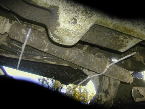 1993 Ford Bronco - Underbody shot looking up at a truck frame rail and crossmember, showing a surface-rusted exhaust pipe, corrugated fuel or brake lines and…