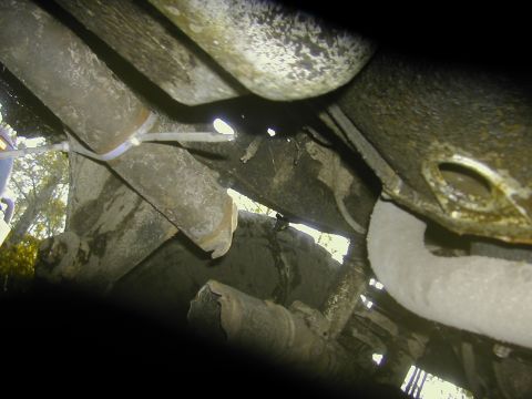 1993 Ford Bronco - Underbody view looking up beneath a 4x4, showing wet, mud-streaked frame rails, a body mount hole, exhaust piping and cable routing secured…