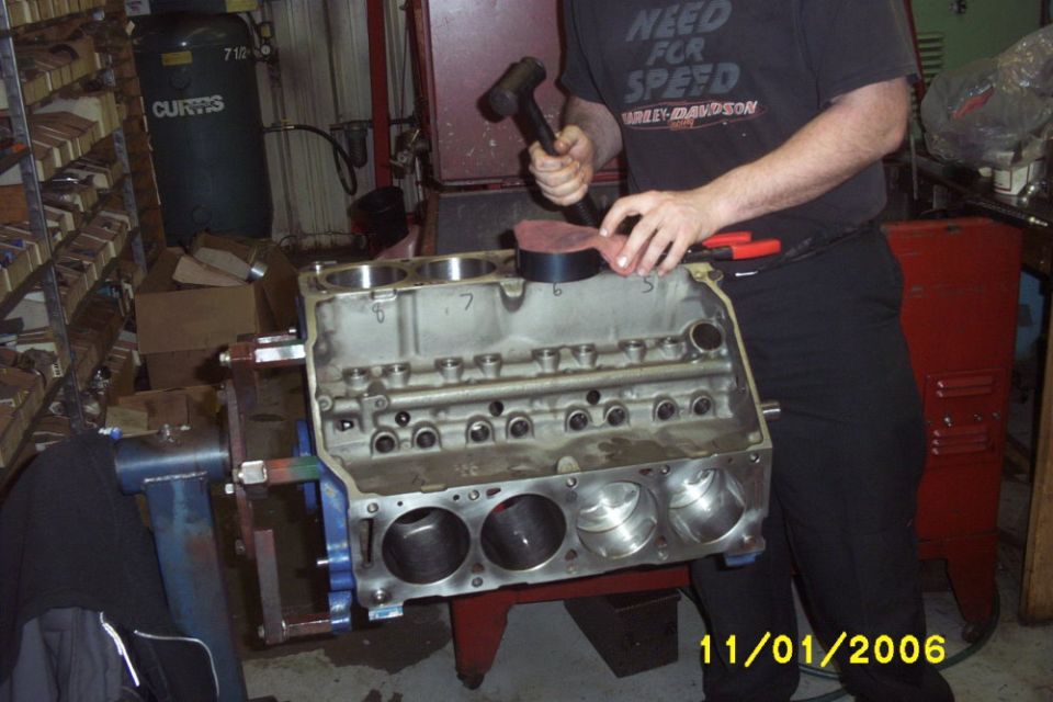 Bare V8 engine block on a stand as a person taps a piston in with a mallet in a shop