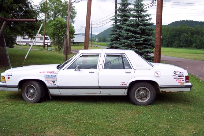 1988 Mercury Grand Marquis sedan with numerous stickers on the doors and rear quarter panel, parked on grass