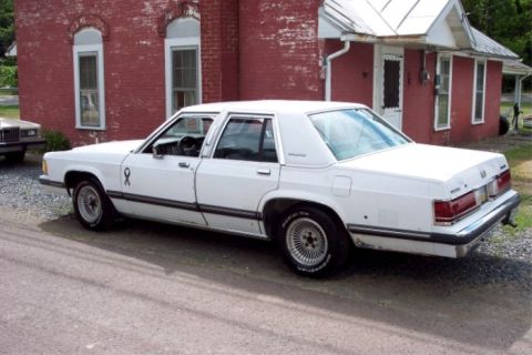 1988 Mercury Grand Marquis - A white sedan is parked in front of a red brick building with white trim.