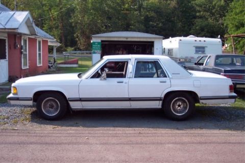 1988 Mercury Grand Marquis - This white sedan is parked next to a garage and a trailer, with another vehicle visible behind it.