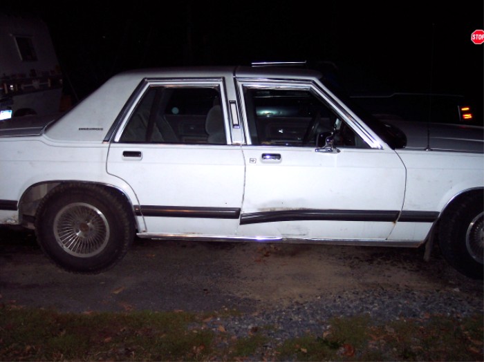 White sedan with chrome trim, dented driver's side door, and wire-spoke wheels, viewed from the side