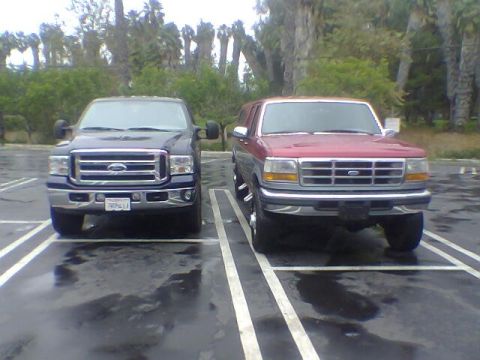 1988 Ford Bronco - Two Ford SUVs are parked side-by-side in a wet parking lot with trees in the background.
