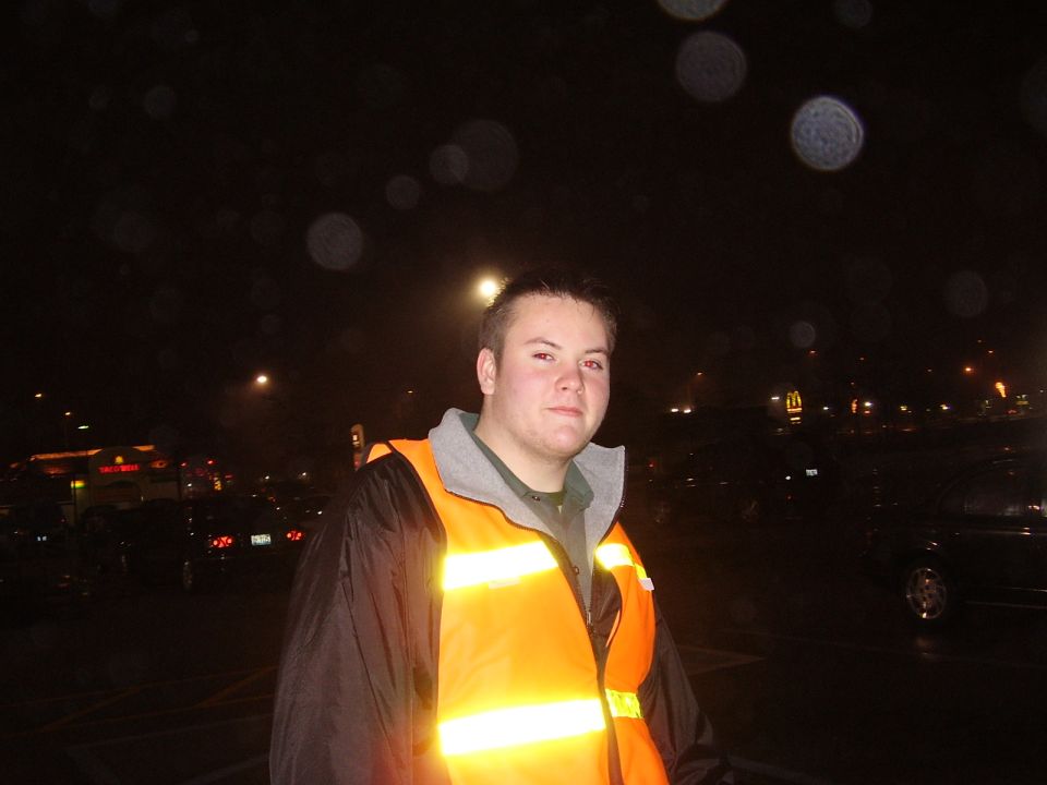Young man in an orange reflective safety vest standing in a dark rainy parking lot