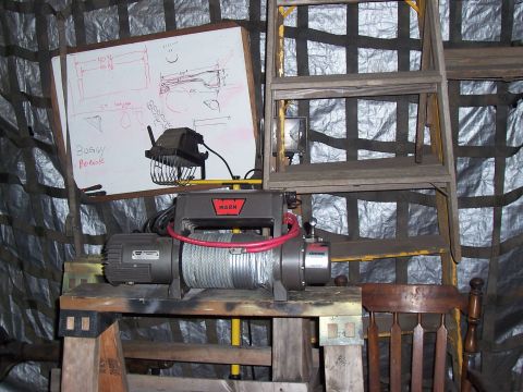 1988 Suzuki Samurai - A Warn winch is displayed on a stand next to a whiteboard with diagrams and text, with a wooden ladder in the background.