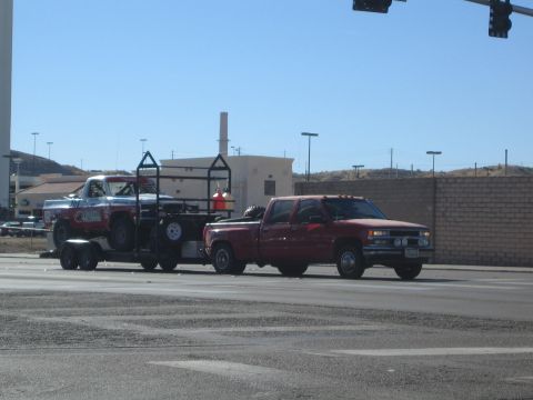 1990 Ford Bronco - red crew-cab dually pickup tows a flatbed trailer carrying a race truck through a road intersection.